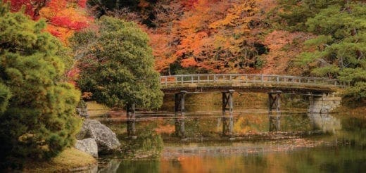 A wooden bridge crosses a calm body of water, surrounded by trees displaying autumn colors.