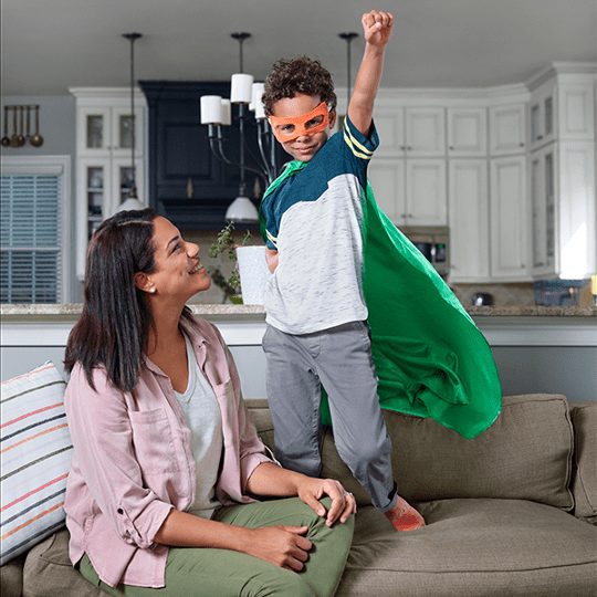 A woman smiles at a child in a superhero costume with a raised fist.