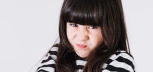 A young girl with dark hair and bangs scrunches her face and crosses her arms.