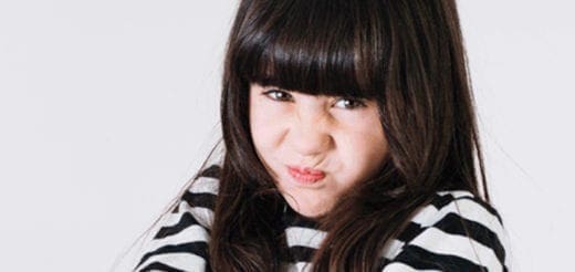 A young girl with dark hair and bangs scrunches her face and crosses her arms.
