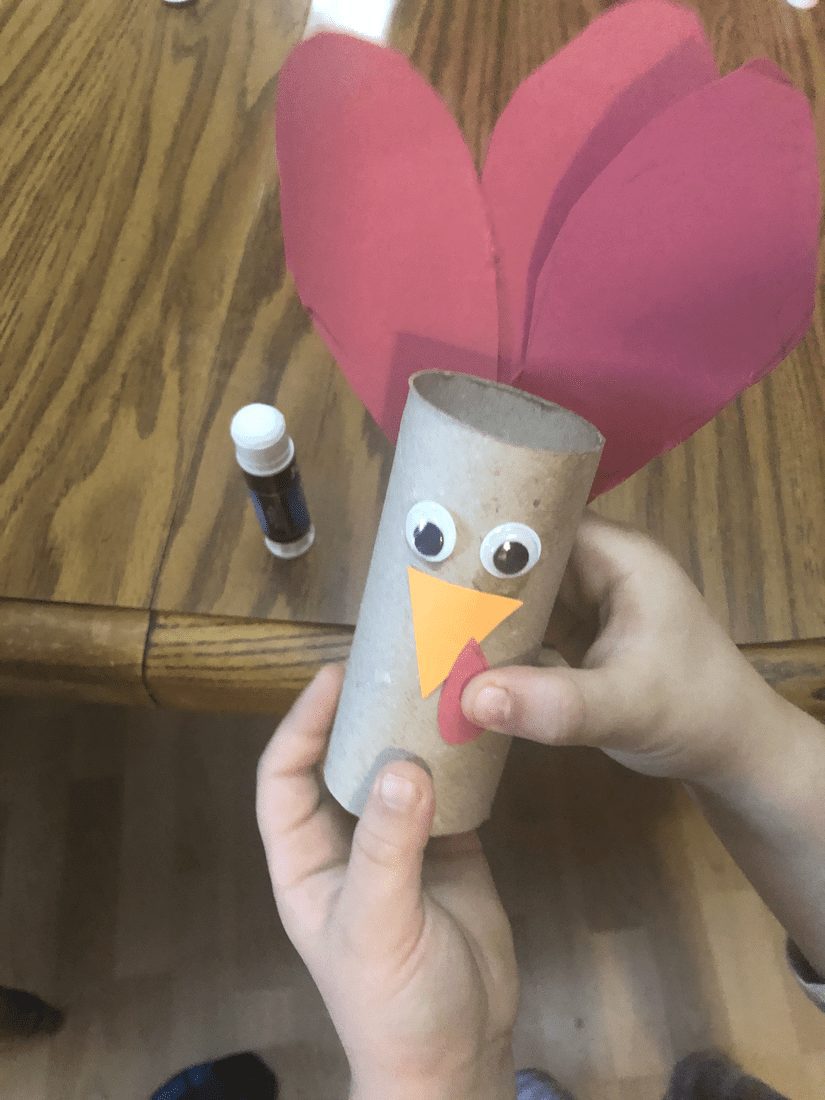 Hands assembling a turkey craft made from a toilet paper tube with googly eyes and a paper beak and wattle.