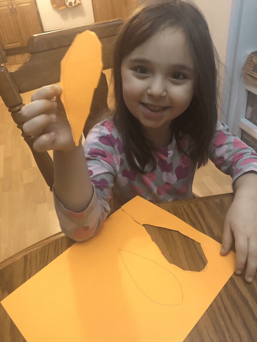 Young girl holds up a cut-out orange feather shape next to an orange paper with a feather outline.