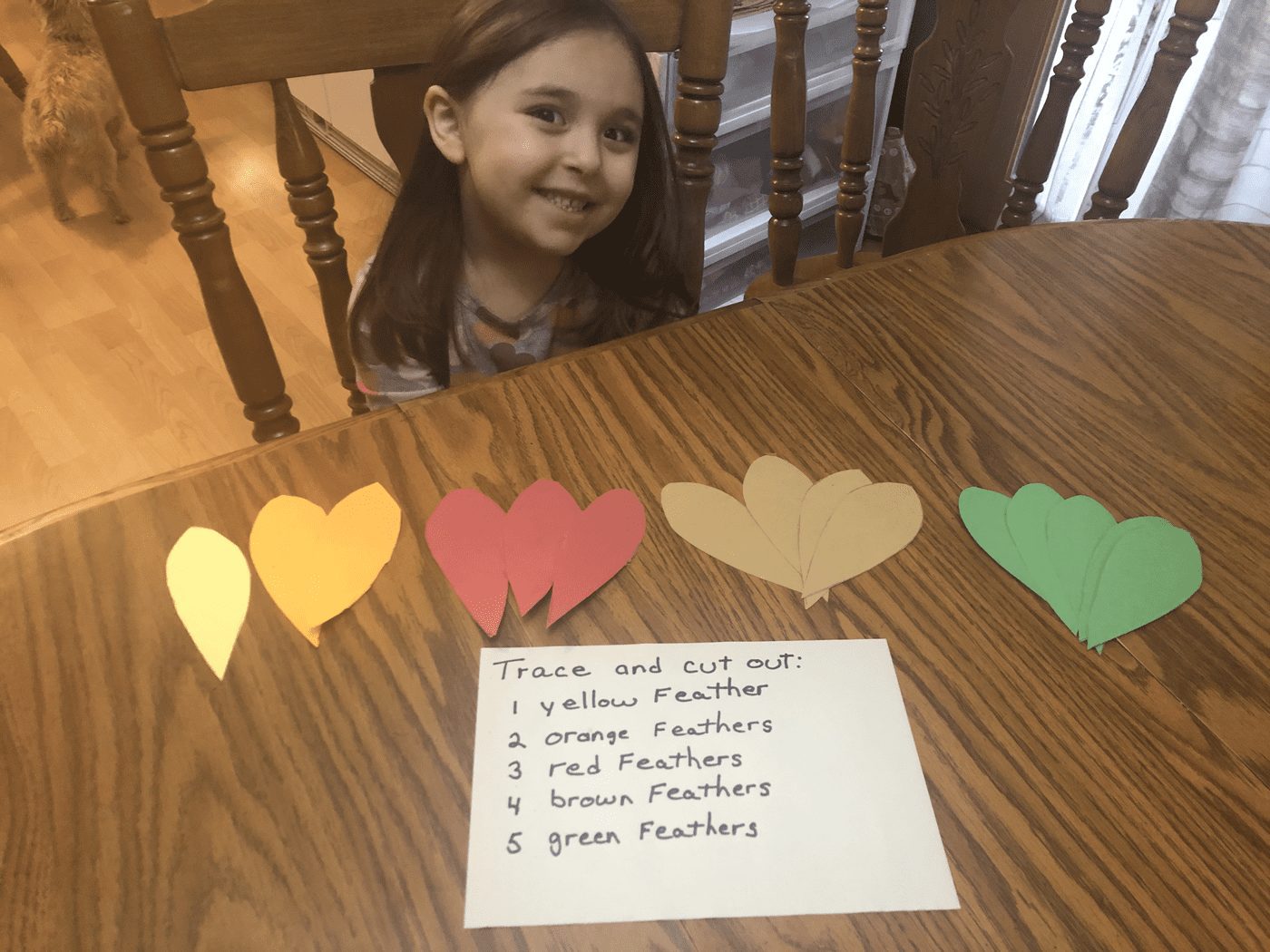 A young girl smiles behind a table with five paper feathers and a list of instructions.