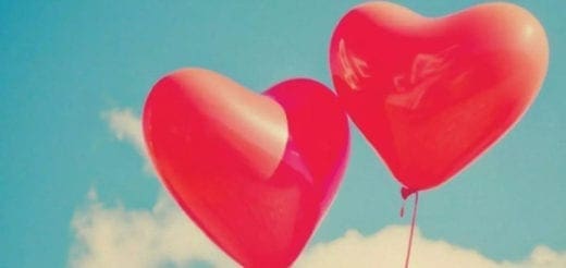 Two red heart-shaped balloons float against a blue sky with white clouds.