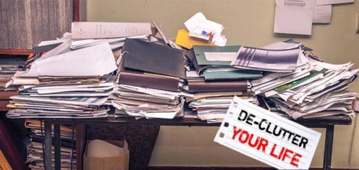 Desk covered in stacks of paper and folders with a sign that says "DE-CLUTTER YOUR LIFE