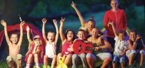 A group of smiling children with their arms raised, gathered around a red guitar in front of a tent.