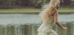 Woman with long blond hair in a white dress running near water.