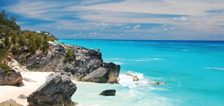 Turquoise water laps against a rocky shore and white sand beach under a blue sky.