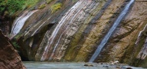 Waterfall cascading down a colorful, layered rock face into a river in Zion National Park.