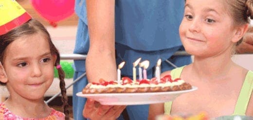 Birthday cake with lit candles being held by an adult, with two young girls watching.