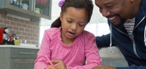 A father smiles as he helps his daughter with her homework at a kitchen table.