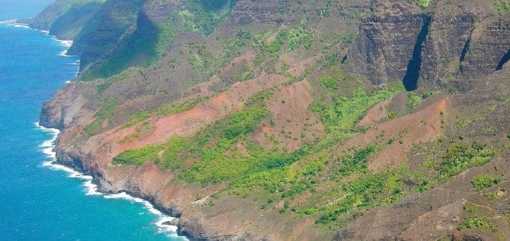 The Na Pali Coast of Kauai, Hawaii, with lush green vegetation on steep, reddish-brown cliffs meeting the turquoise ocean.
