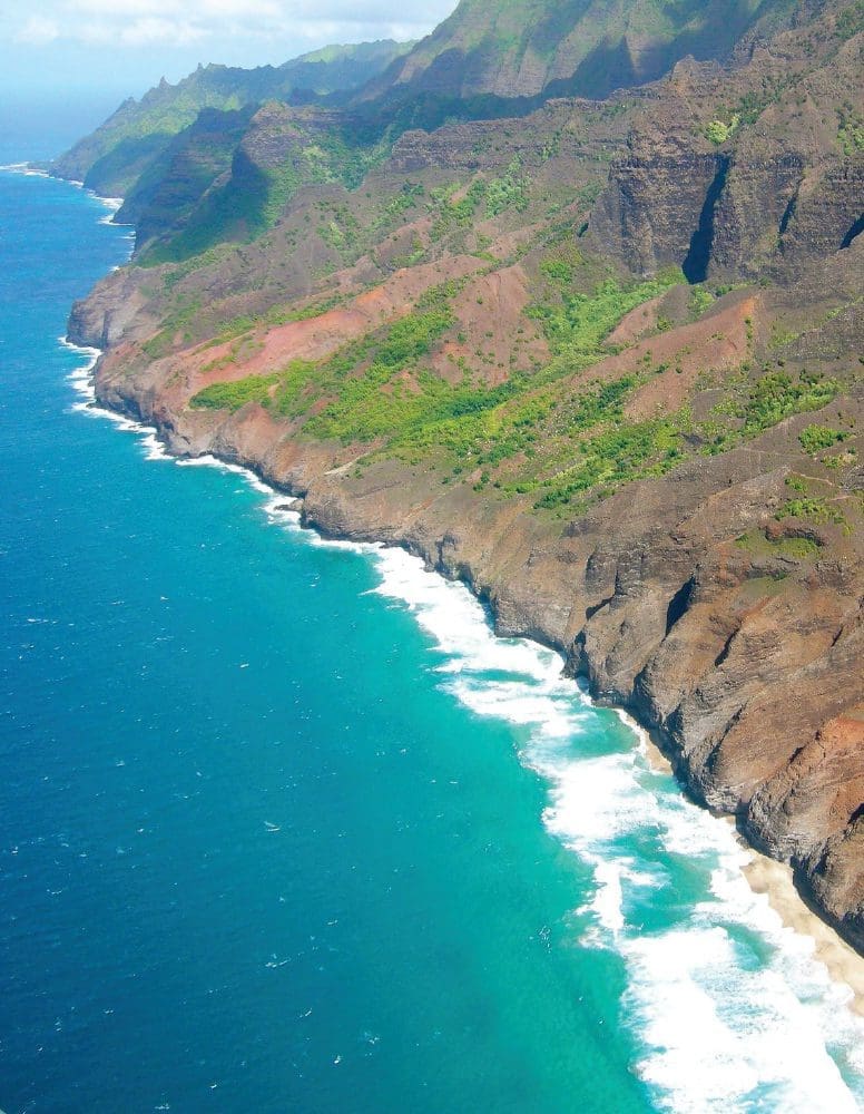 NaPali Coast, Kauai, with turquoise ocean waves crashing on the shore below steep, green, and brown cliffs.