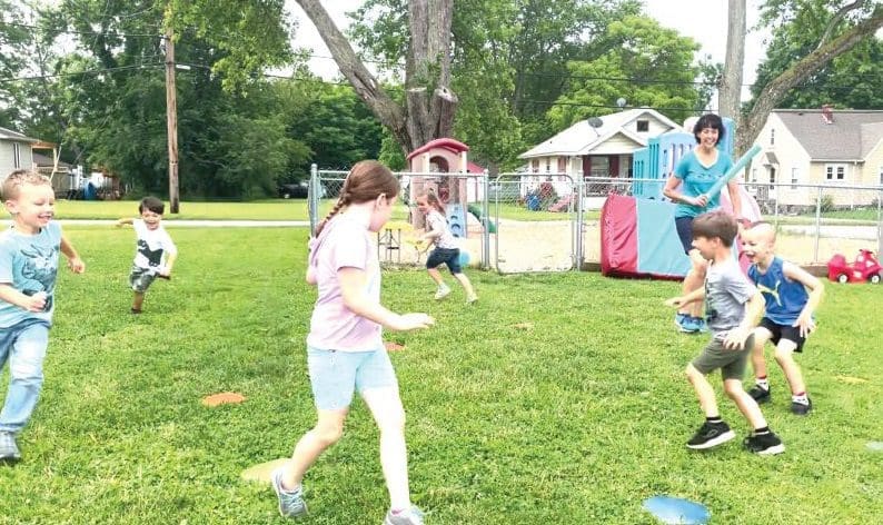Children play a game on a grassy lawn with a playground in the background.