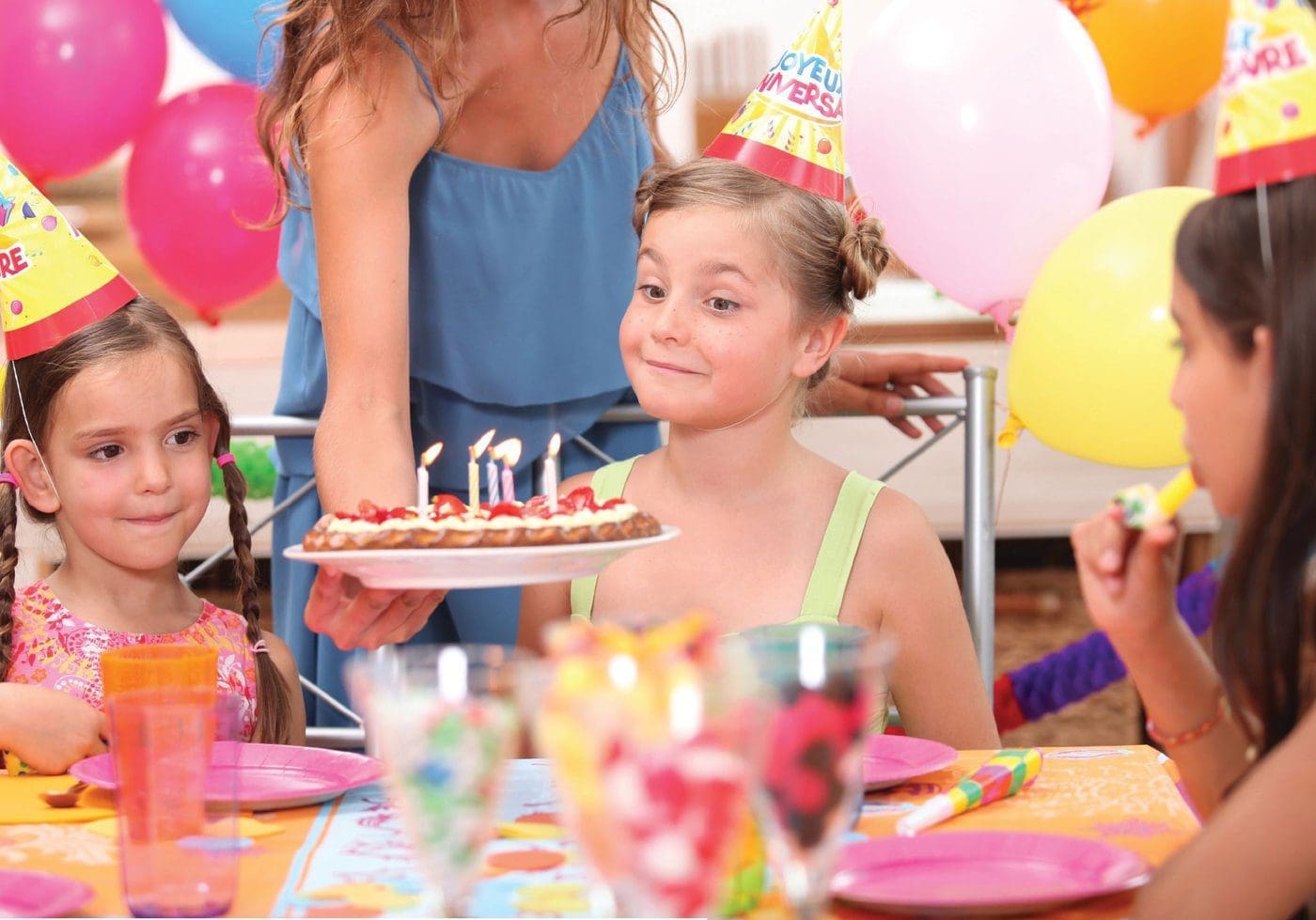Children in party hats look at a cake with lit candles held by an adult.