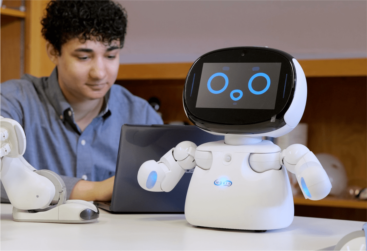 A white robot with a screen face stands on a table in front of a boy using a laptop.