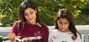 Two young women study outdoors, one holding a book and pencil.