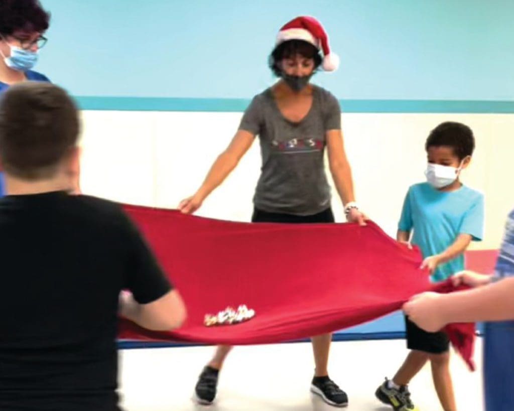 Four people wearing masks hold a red sheet with bells on it, one person wears a Santa hat.