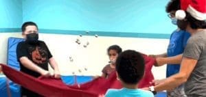 Children in masks holding a red cloth with a few objects in the air.
