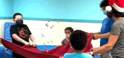 Children in masks holding a red cloth with a few objects in the air.