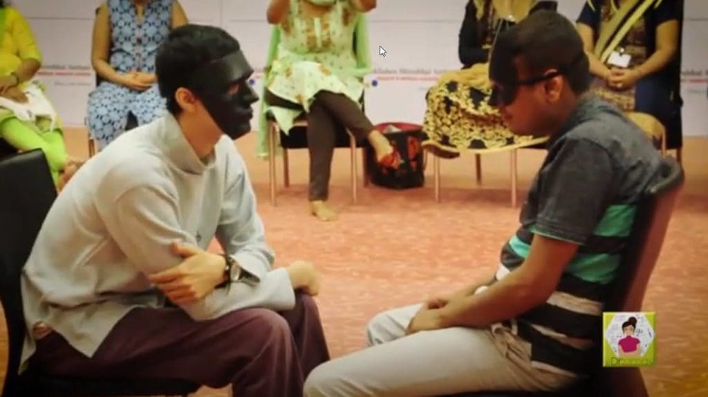 Two men sit facing each other, one wearing a black mask, with an audience seated in the background.