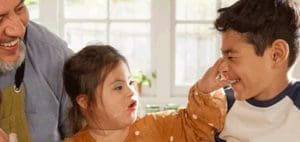 A smiling boy teases a girl with Down syndrome while a man looks on.