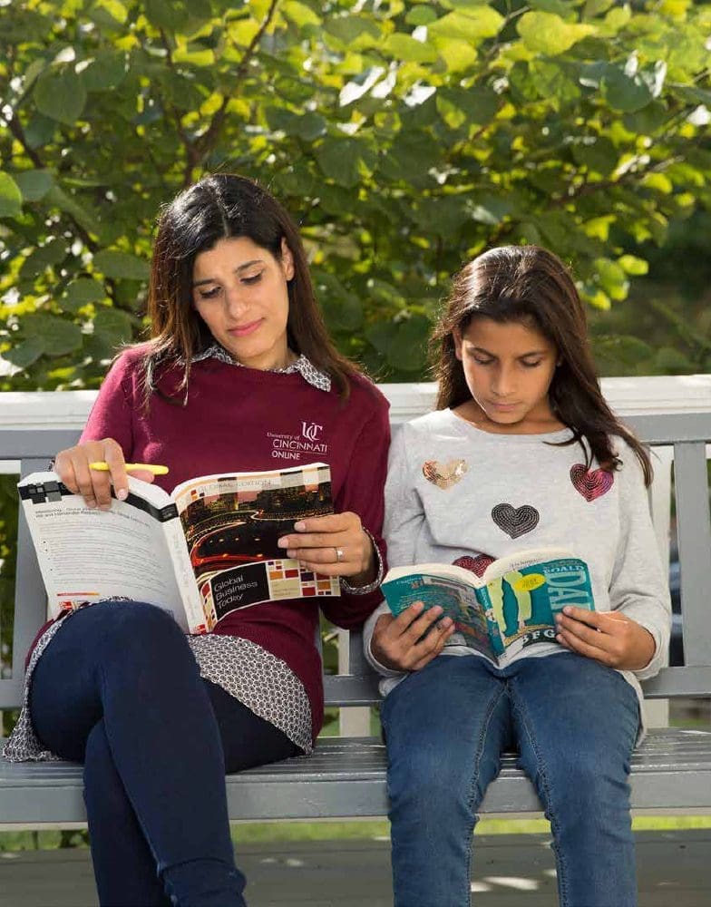 Two women sitting on a bench outdoors reading books.