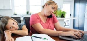 A child rests their head on their arms while a woman on the phone types on a laptop.