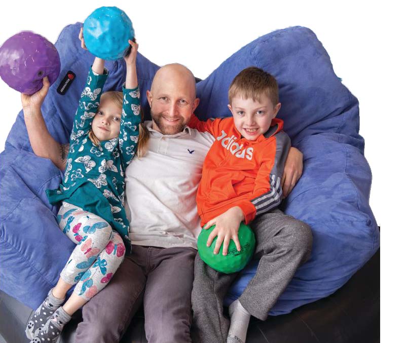 A man sits on a large blue beanbag chair with two children, each holding a colorful ball.