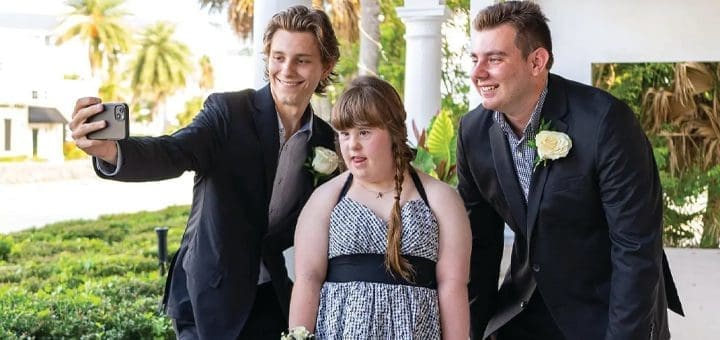 Three people take a selfie outdoors, two men in suits flanking a young woman in a dress.