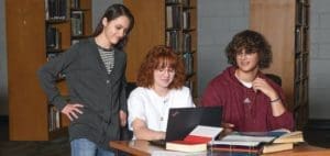 Three students collaborate on a laptop in a library setting.