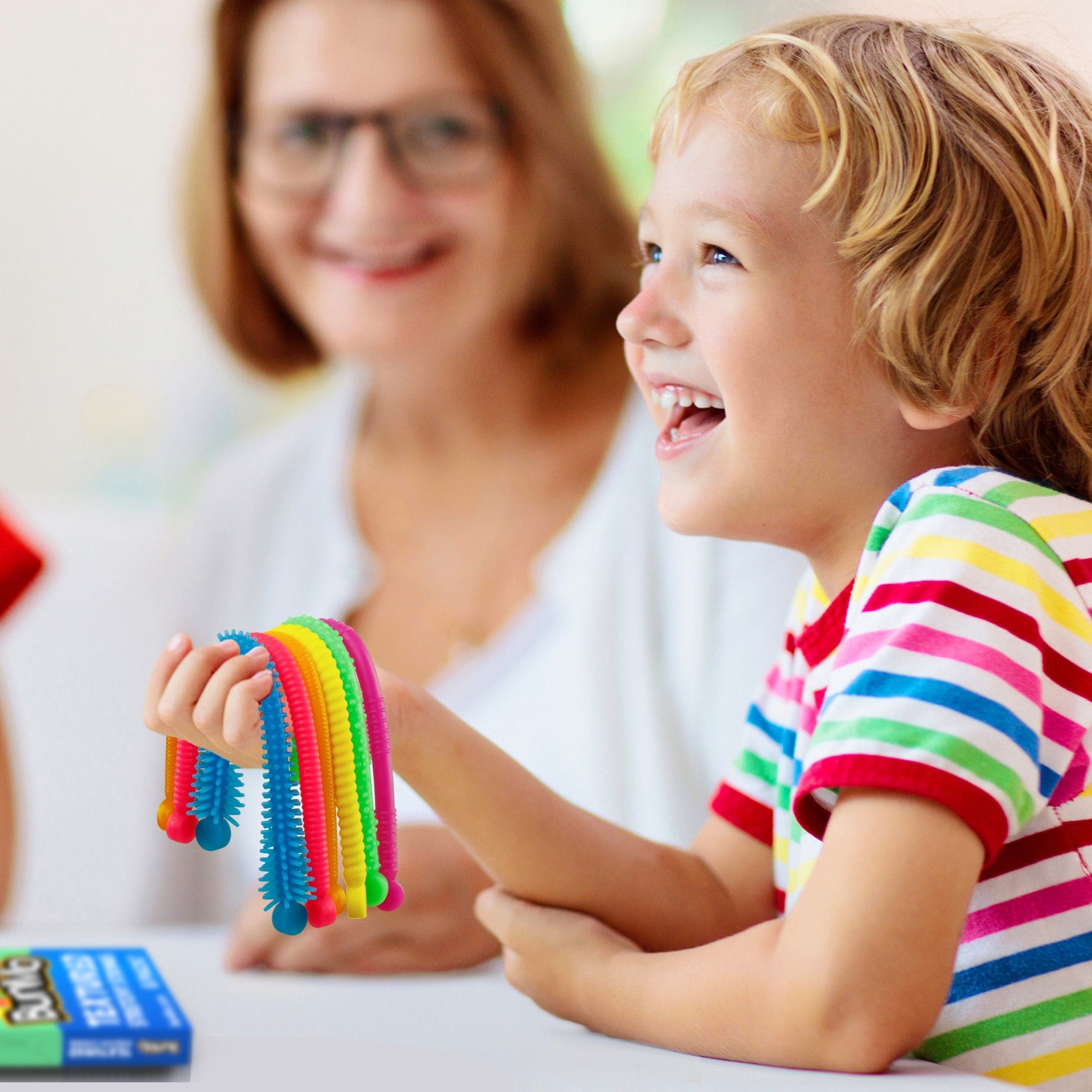 A child smiles while holding a rainbow-colored BunMo fidget toy, with an adult blurred in the background.