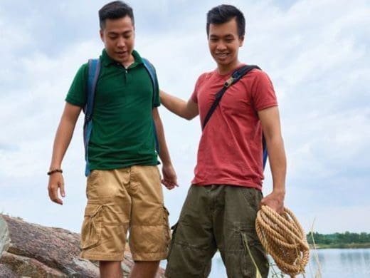 Two men with backpacks and a rope stand outdoors.