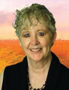 A woman smiles at the camera, wearing a black top and a beaded necklace, with a sunset-like background.