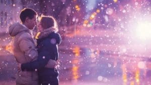 Couple embracing in falling snow at night with blurred city lights in the background.