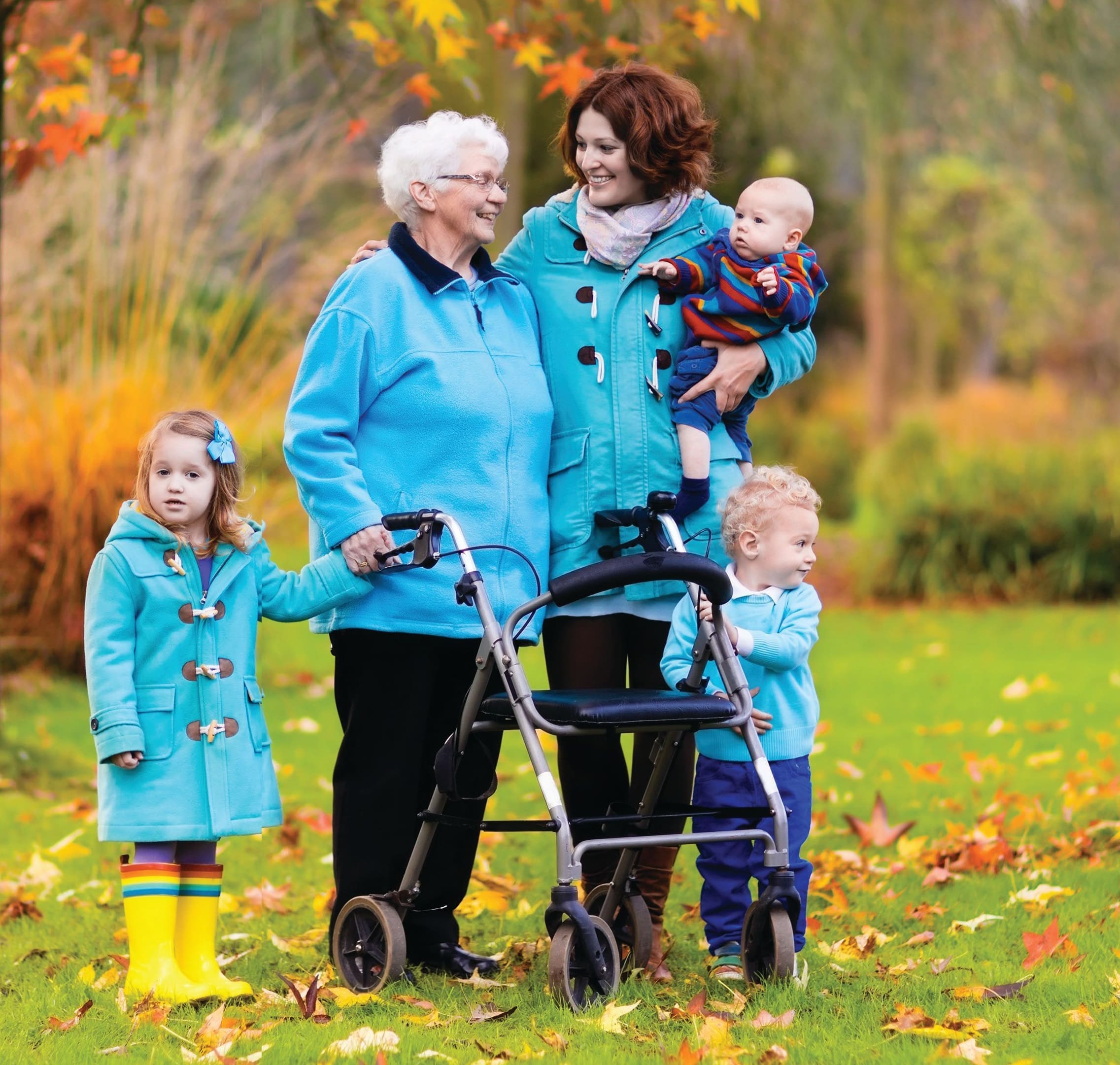 Family with a baby, toddler, older child, and elderly woman with a walker in autumn leaves.