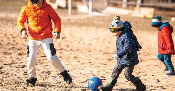 Three children play soccer on a sandy field.