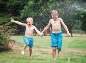 Two young boys in swim trunks run through a sprinkler in a grassy yard.