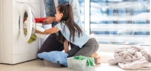 Child loading laundry into a washing machine with adult's help.