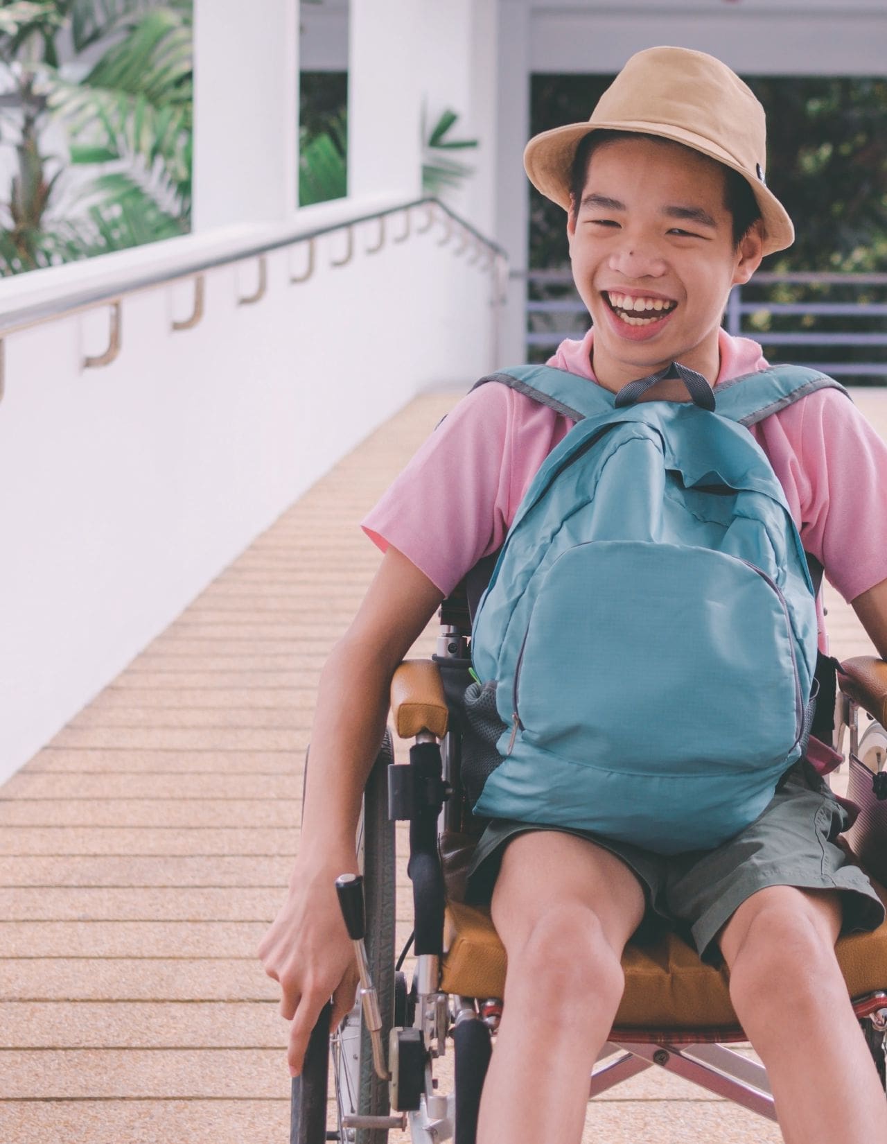 A smiling child in a wheelchair wearing a pink shirt, green shorts, and a hat, with a blue backpack.