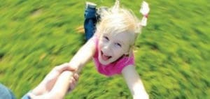 A child with blonde hair held by adult hands, spinning outdoors with a green, blurry background.