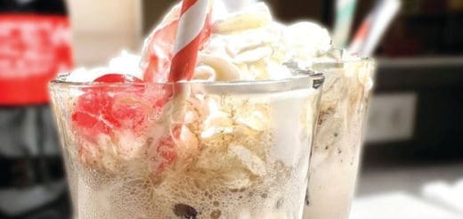 Two ice cream sodas with whipped cream and cherries, one with a striped straw.