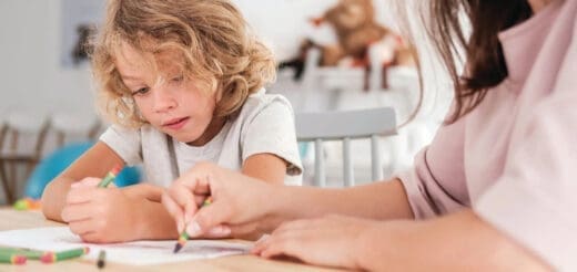 A child with blonde, curly hair draws with a crayon while an adult's hand guides theirs.