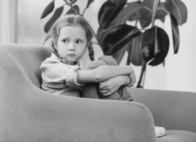 A young girl sits with her arms wrapped around her knees in an armchair.