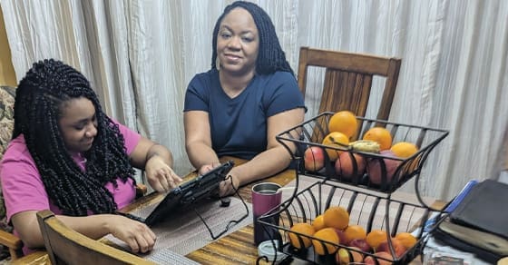 Two women sit at a table with fruit baskets, a tablet, and books.
