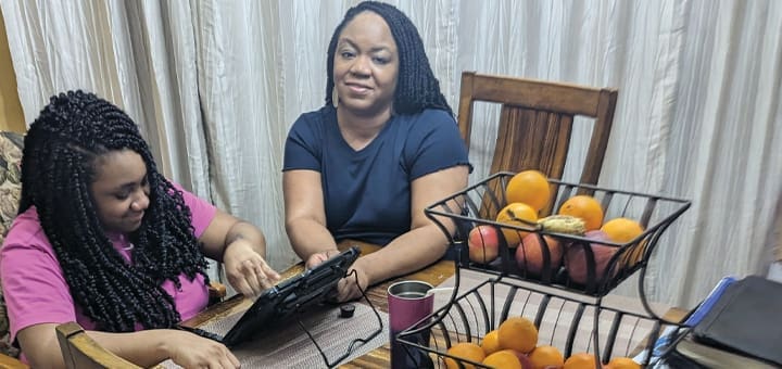Two women sit at a table with a tablet and fruit baskets.