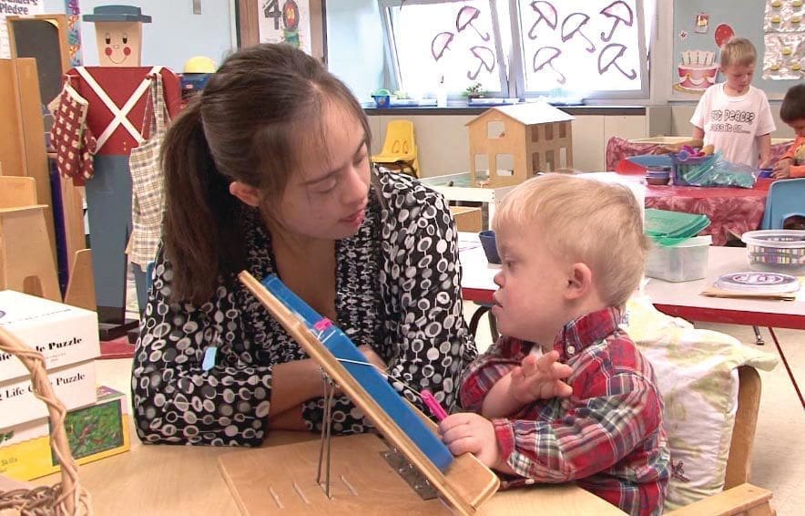 A woman helps a young child with a learning activity at a table.