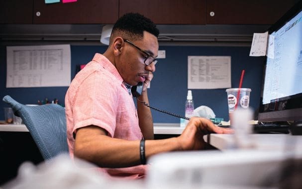 Man in a pink shirt talking on the phone at a desk.