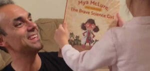 A man smiles as a child holds up the book "Mya McLure, The Brave Science Girl.