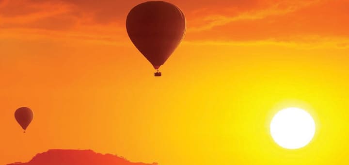 Two hot air balloons float in a bright orange sky above a silhouetted landscape during sunrise.